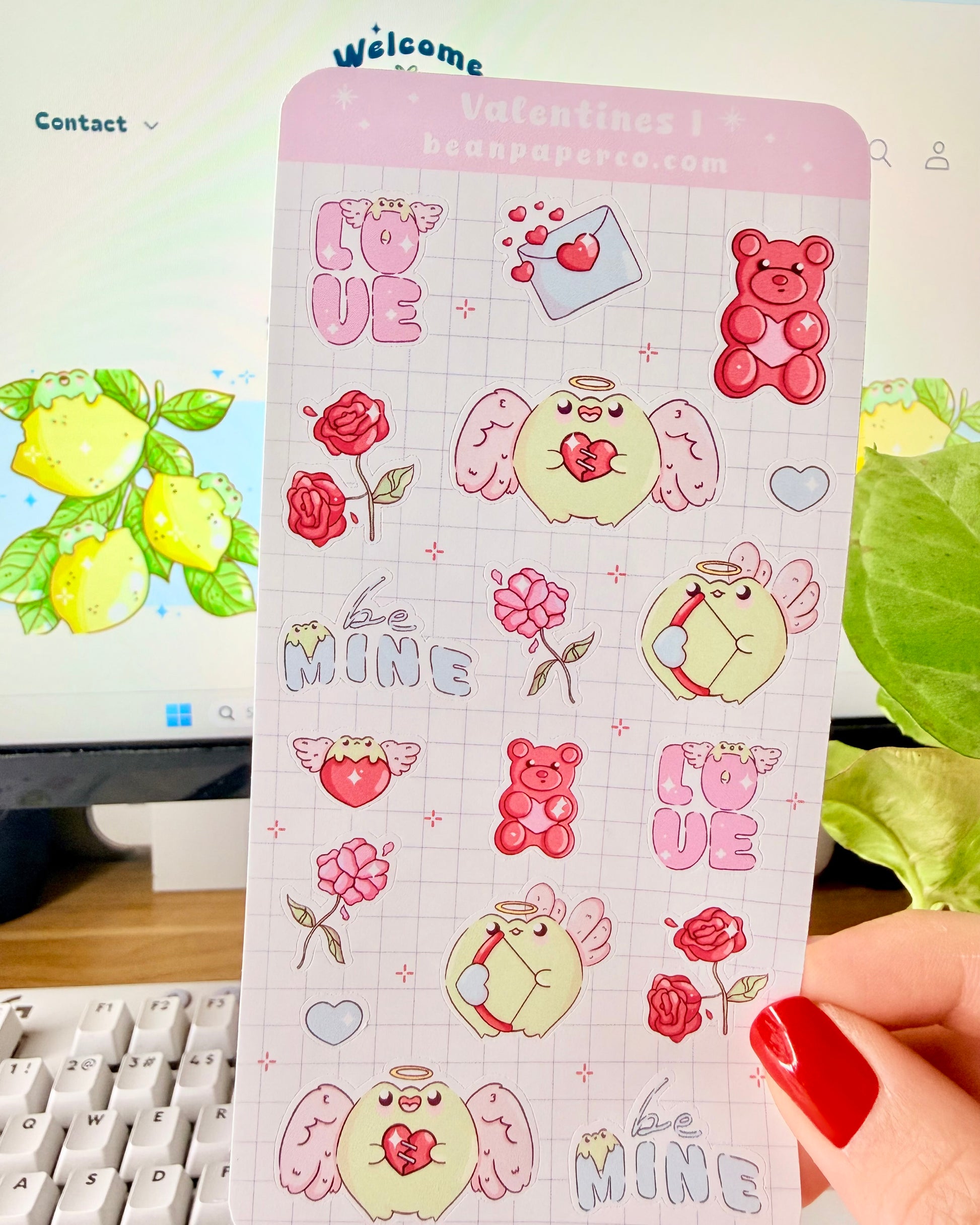 Kawaii Valentine Frog Sticker Sheet. Hand holding a Valentine-themed sticker sheet featuring angel frogs, roses, gummy bears, love letters, and “Be Mine” text in pink and red tones against a computer desk background.
