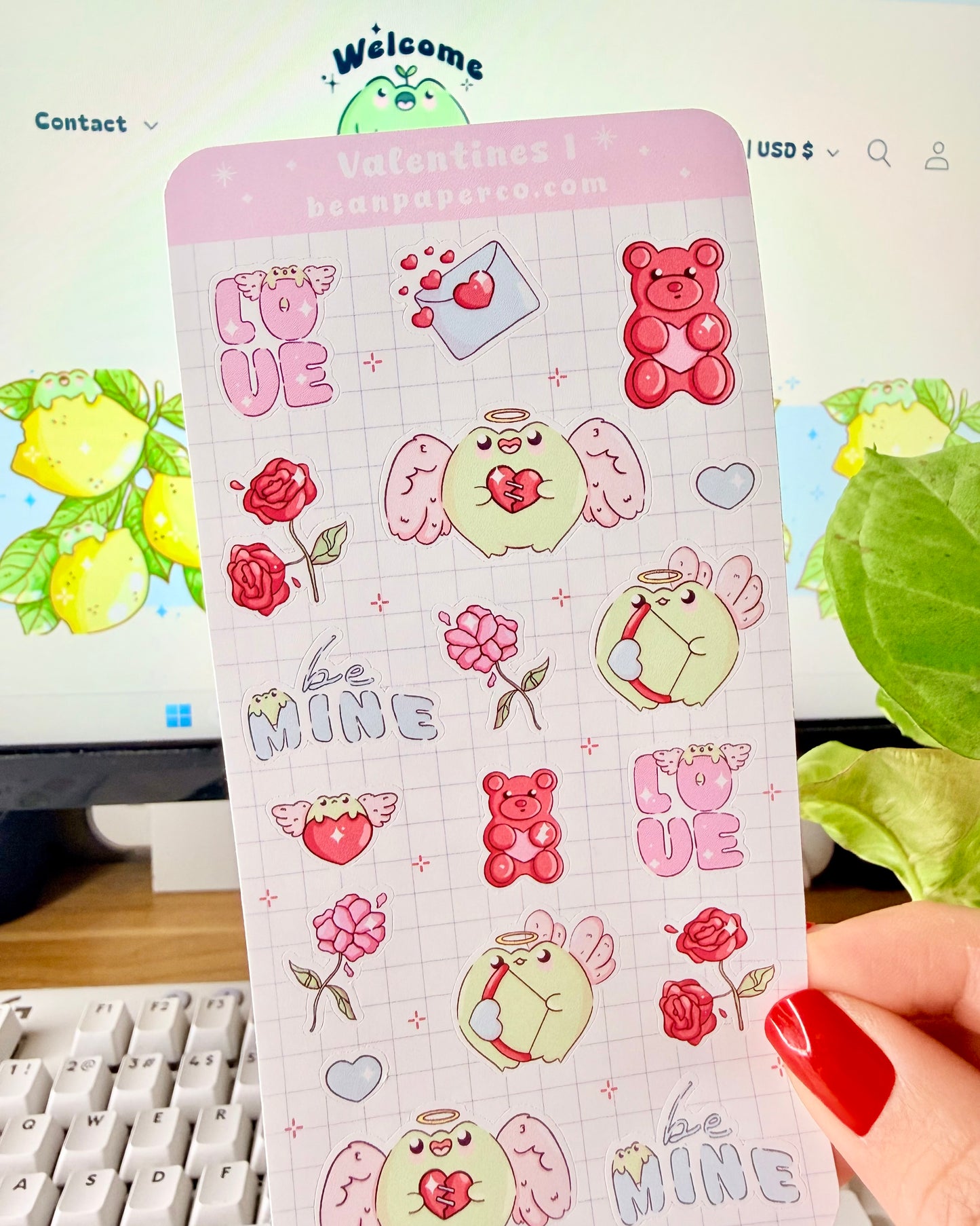 Kawaii Valentine Frog Sticker Sheet. Hand holding a Valentine-themed sticker sheet featuring angel frogs, roses, gummy bears, love letters, and “Be Mine” text in pink and red tones against a computer desk background.