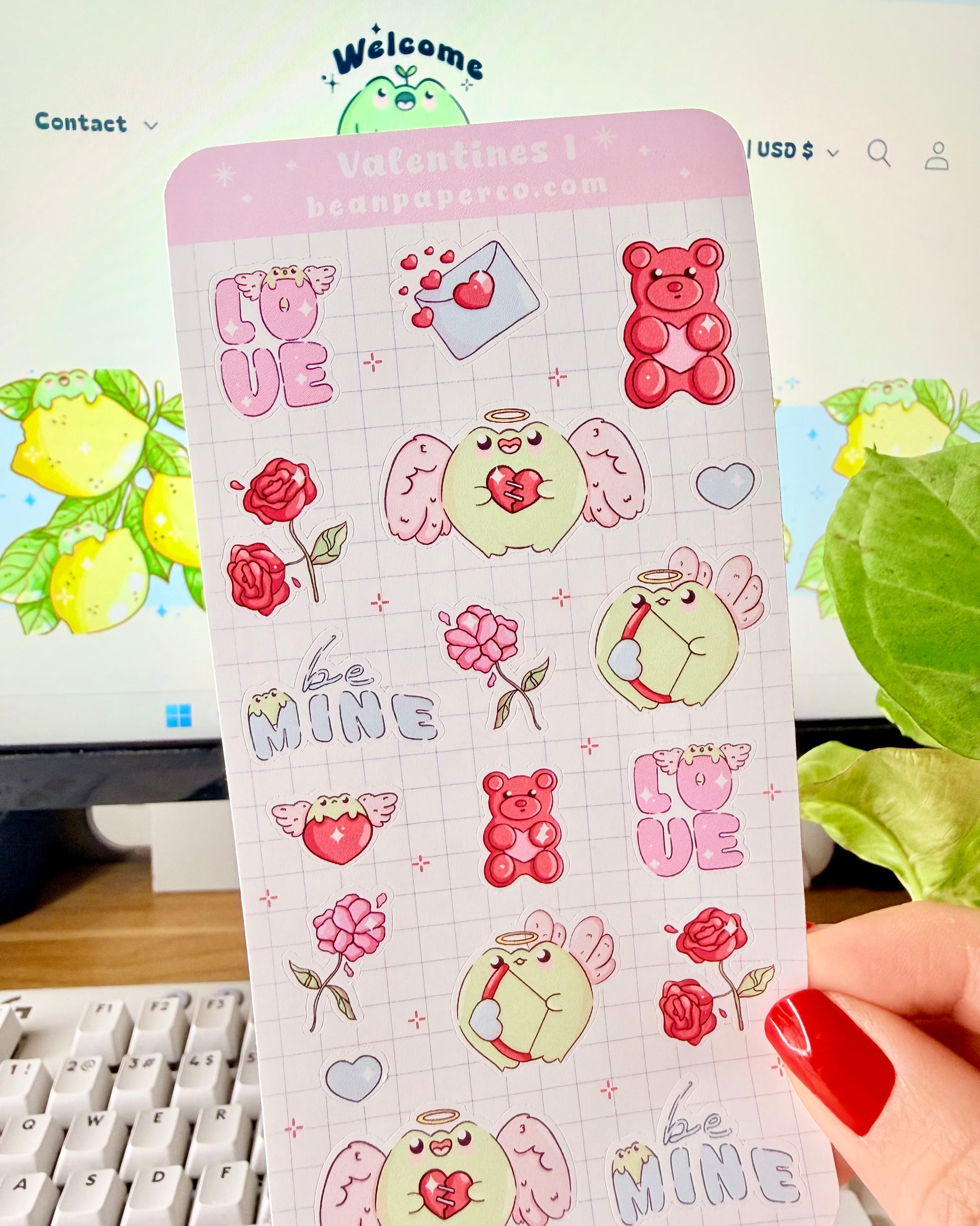 Kawaii Valentine Frog Sticker Sheet. Hand holding a Valentine-themed sticker sheet featuring angel frogs, roses, gummy bears, love letters, and “Be Mine” text in pink and red tones against a computer desk background.
