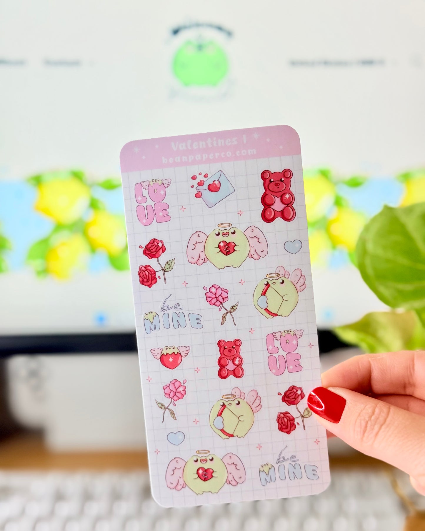 Kawaii Valentine Frog Sticker Sheet. Hand holding a Valentine-themed sticker sheet featuring angel frogs, roses, gummy bears, love letters, and “Be Mine” text in pink and red tones against a computer desk background.