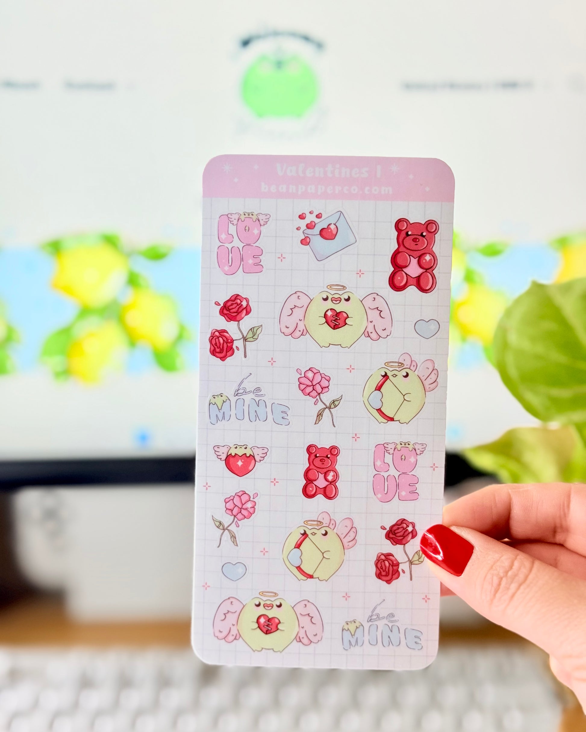 Kawaii Valentine Frog Sticker Sheet. Hand holding a Valentine-themed sticker sheet featuring angel frogs, roses, gummy bears, love letters, and “Be Mine” text in pink and red tones against a computer desk background.