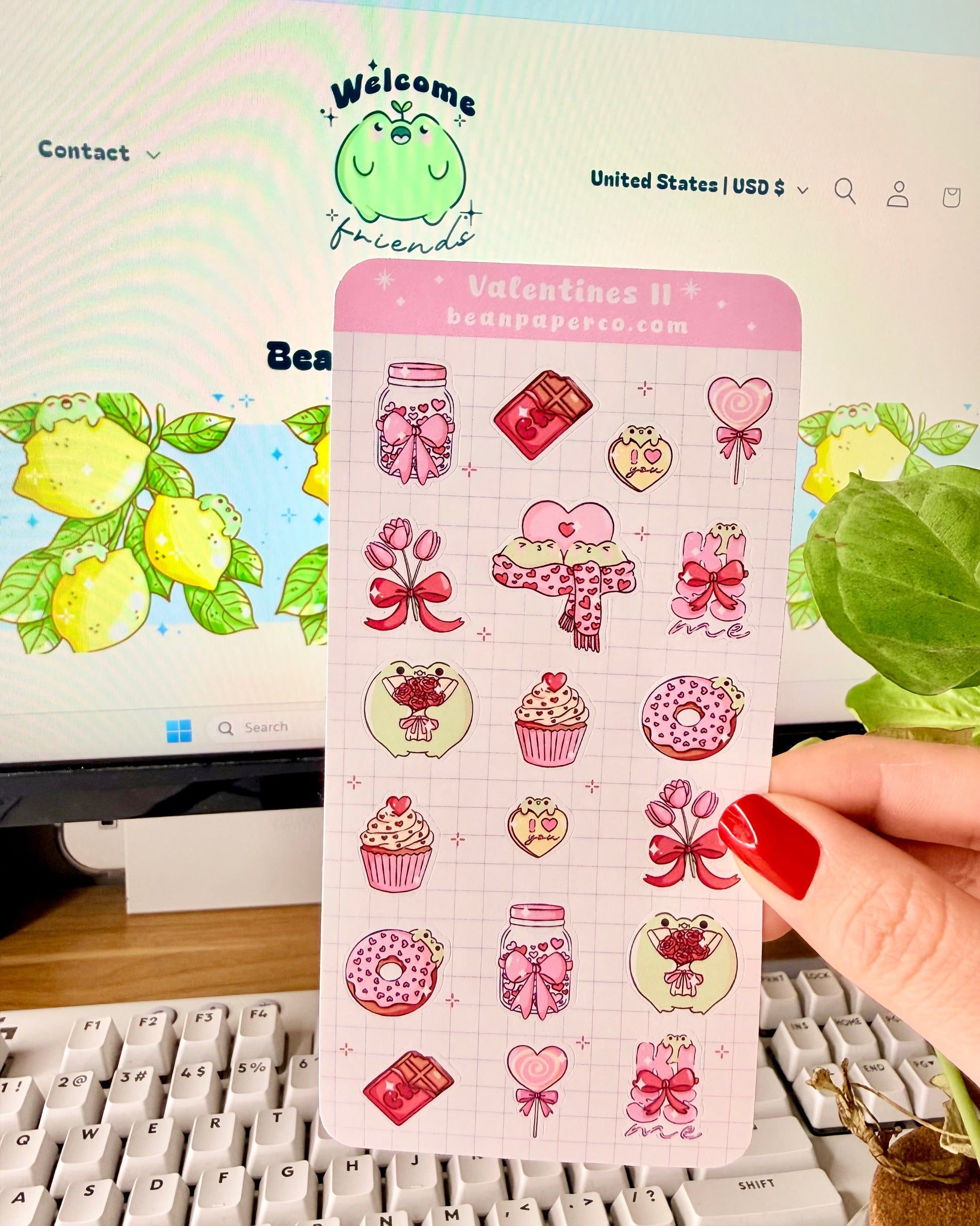 Valentine Frog Dessert Sticker Sheet. Hand holding a pastel Valentine’s sticker sheet featuring pink and red themed designs including cupcakes, donuts, chocolate bars, flowers, heart lollipops, bows, and jars, photographed in front of a computer screen and plant.
