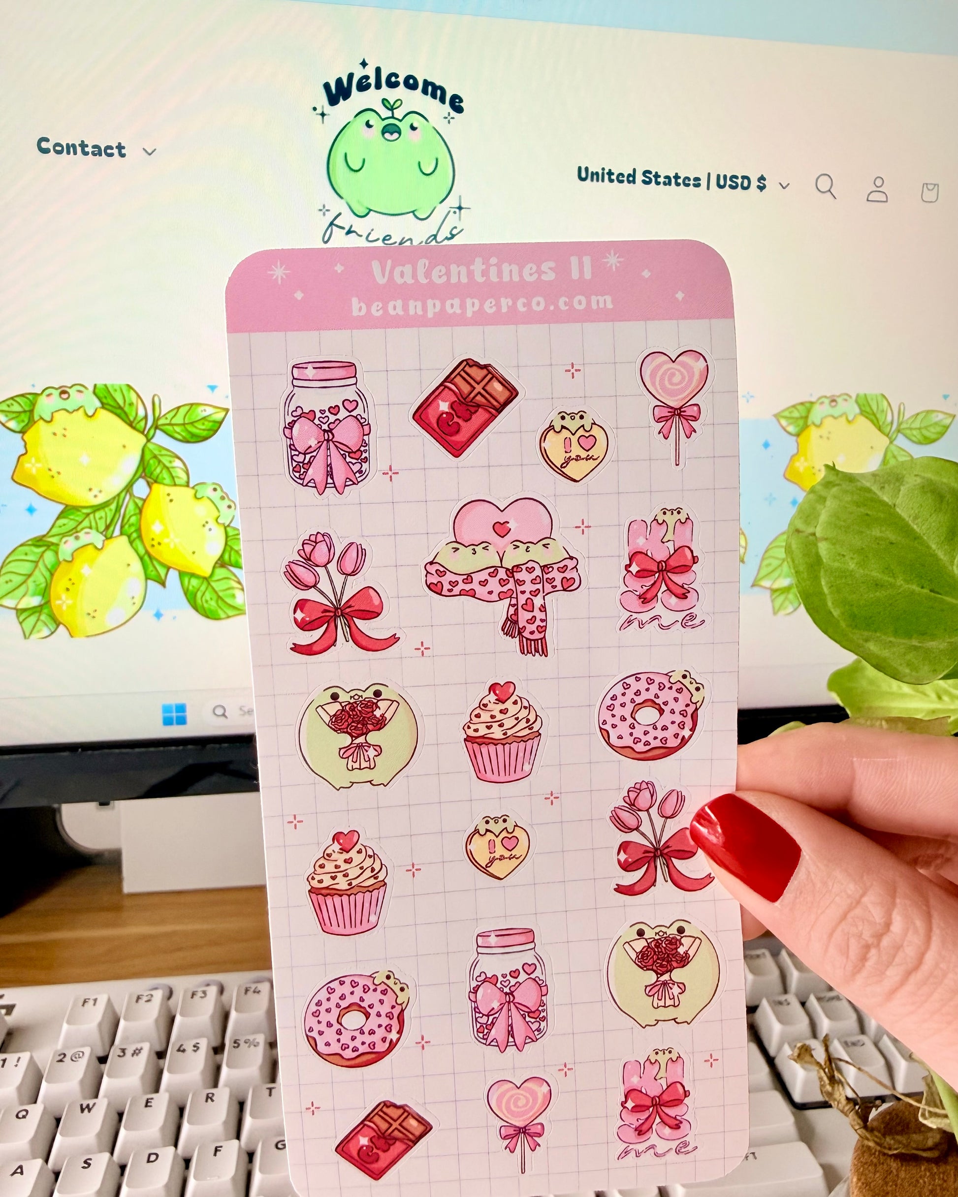 Valentine Frog Dessert Sticker Sheet. Hand holding a pastel Valentine’s sticker sheet featuring pink and red themed designs including cupcakes, donuts, chocolate bars, flowers, heart lollipops, bows, and jars, photographed in front of a computer screen and plant.
