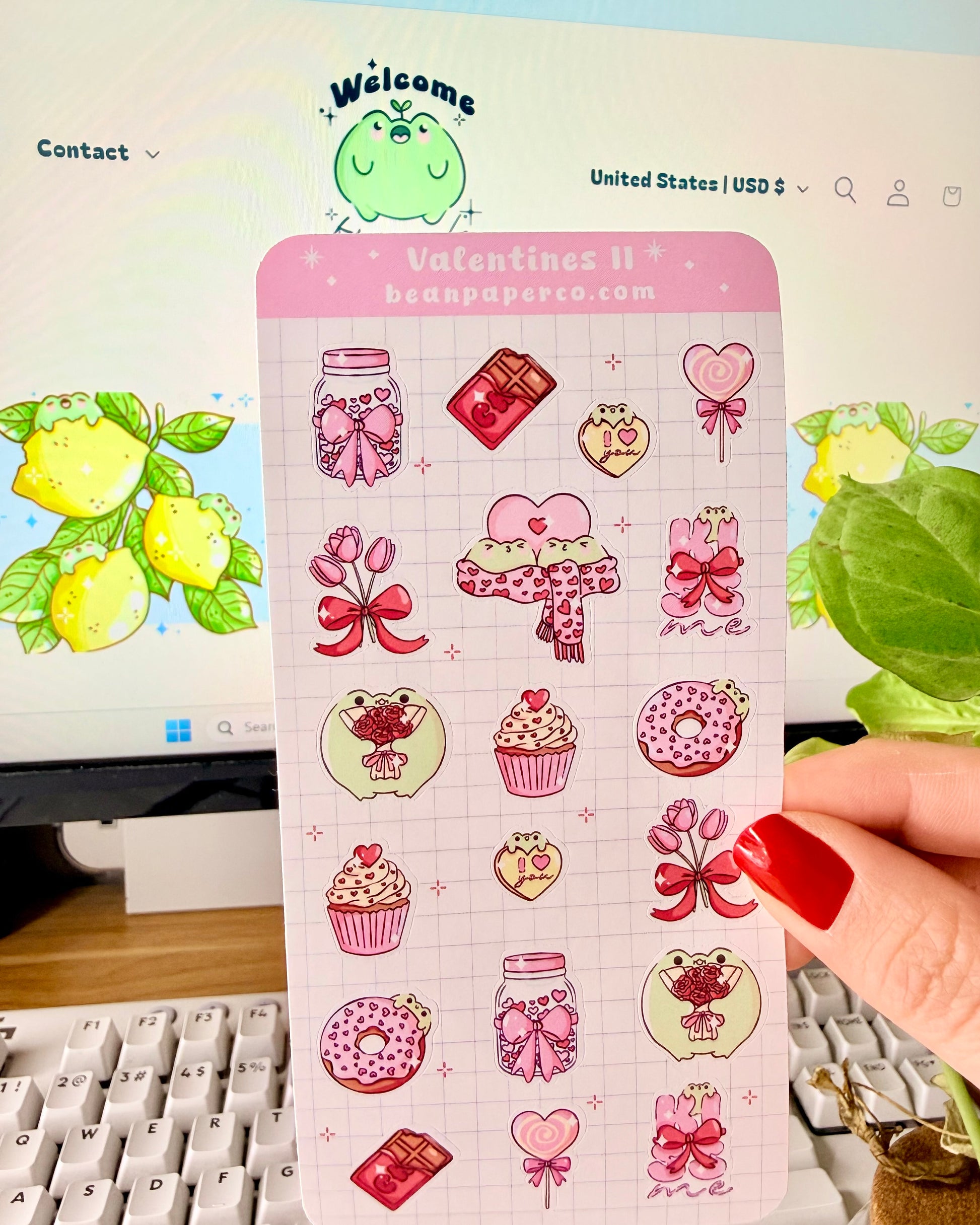 Valentine Frog Dessert Sticker Sheet. Hand holding a pastel Valentine’s sticker sheet featuring pink and red themed designs including cupcakes, donuts, chocolate bars, flowers, heart lollipops, bows, and jars, photographed in front of a computer screen and plant.