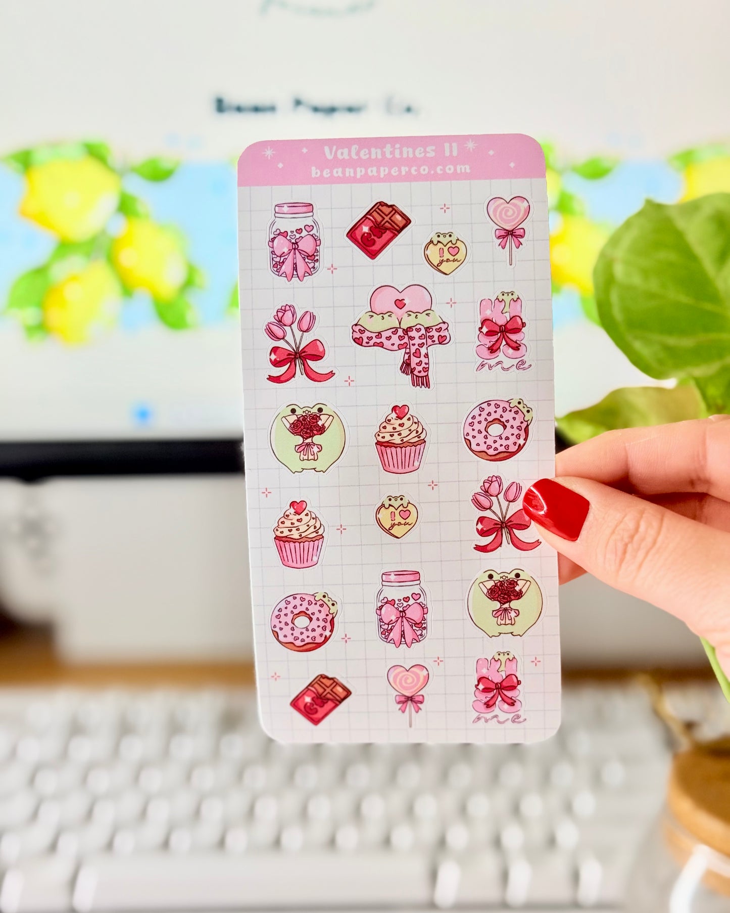 Valentine Frog Dessert Sticker Sheet. Hand holding a pastel Valentine’s sticker sheet featuring pink and red themed designs including cupcakes, donuts, chocolate bars, flowers, heart lollipops, bows, and jars, photographed in front of a computer screen and plant.