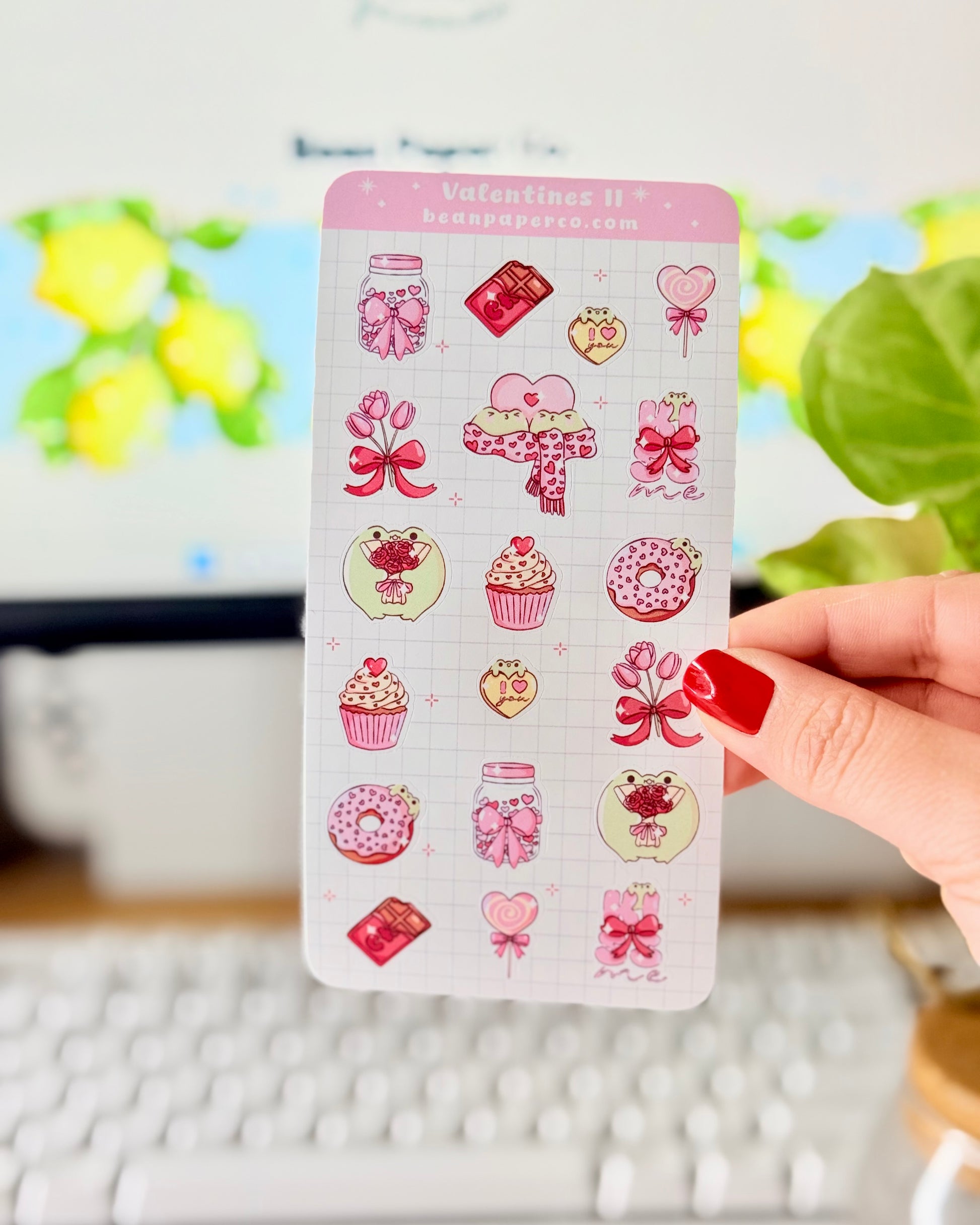 Valentine Frog Dessert Sticker Sheet. Hand holding a pastel Valentine’s sticker sheet featuring pink and red themed designs including cupcakes, donuts, chocolate bars, flowers, heart lollipops, bows, and jars, photographed in front of a computer screen and plant.