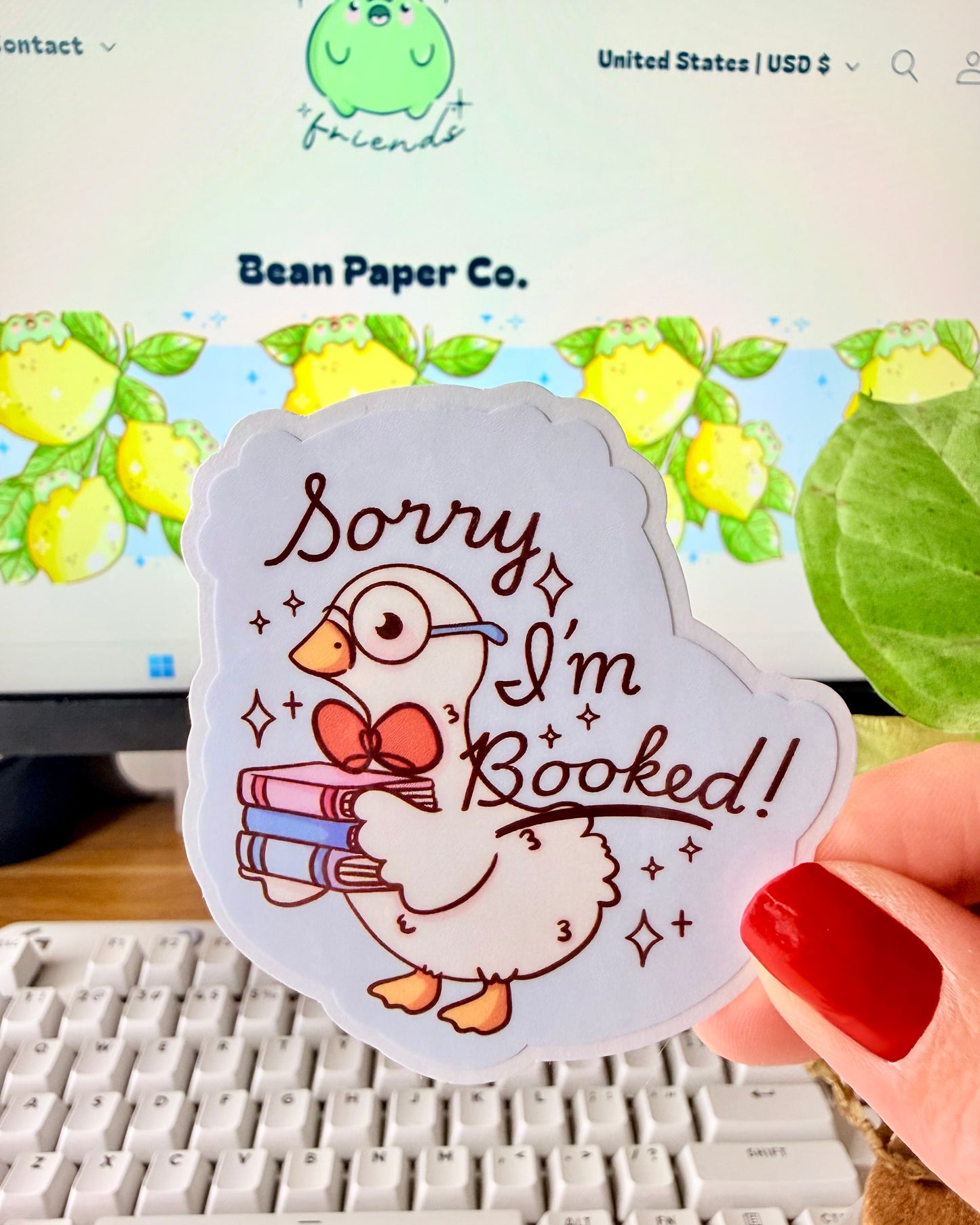 Adorable I'm Booked Duck Sticker. Hand holding a pastel vinyl sticker of a white goose wearing glasses and holding a stack of books with the text “Sorry I’m Booked!” in front of a computer keyboard and plant.