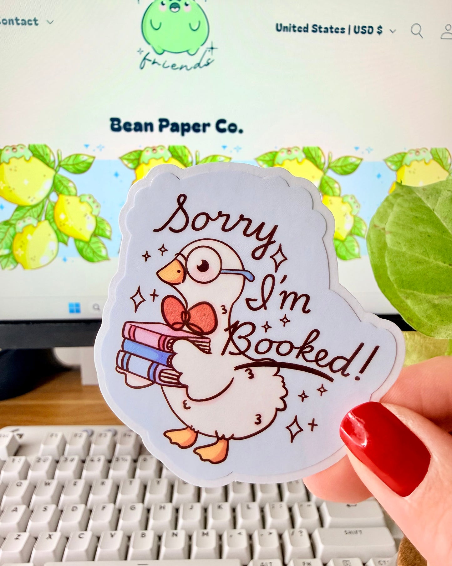 Adorable I'm Booked Duck Sticker. Hand holding a pastel vinyl sticker of a white goose wearing glasses and holding a stack of books with the text “Sorry I’m Booked!” in front of a computer keyboard and plant.
