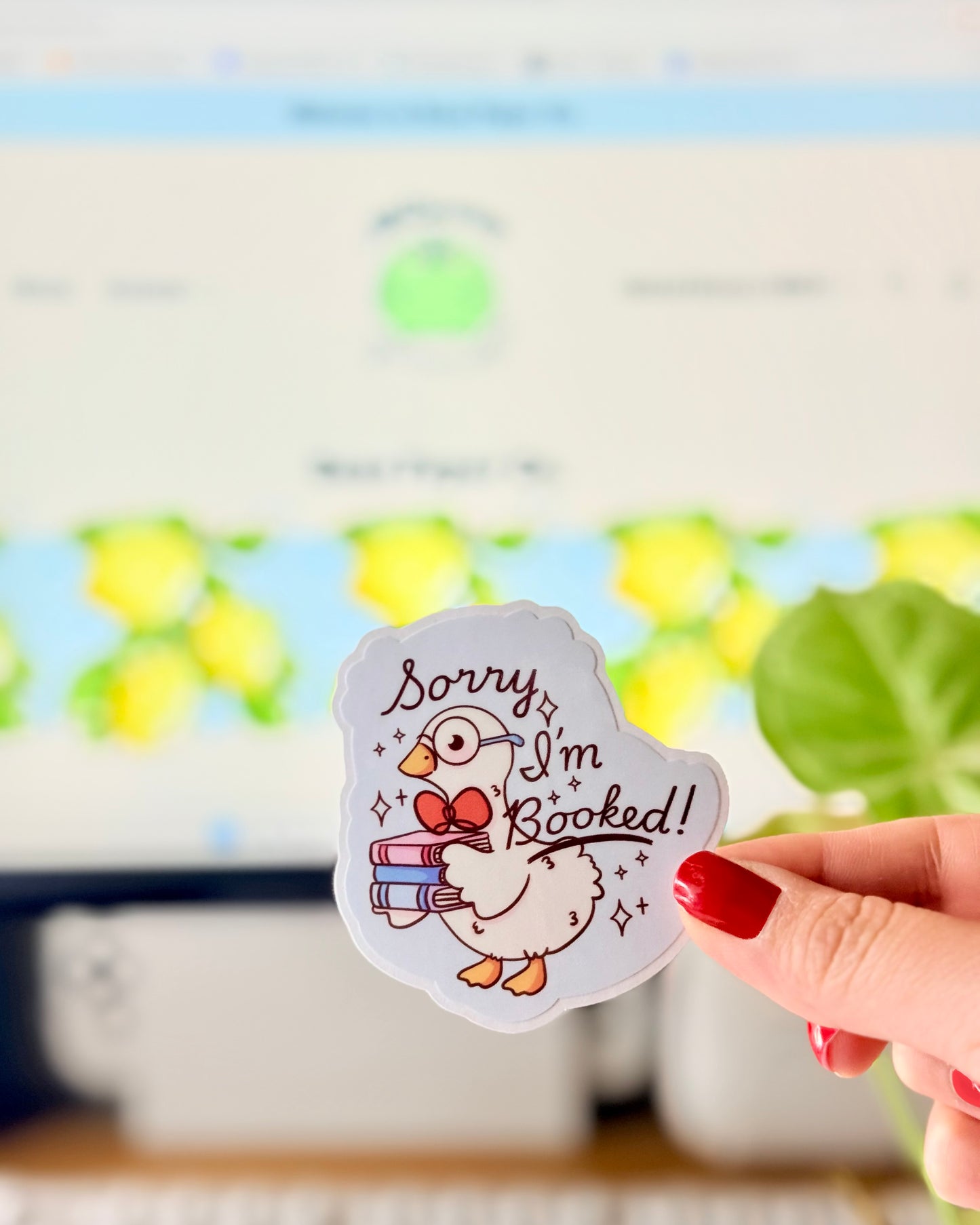 Adorable I'm Booked Duck Sticker. Hand holding a pastel vinyl sticker of a white goose wearing glasses and holding a stack of books with the text “Sorry I’m Booked!” in front of a computer keyboard and plant.