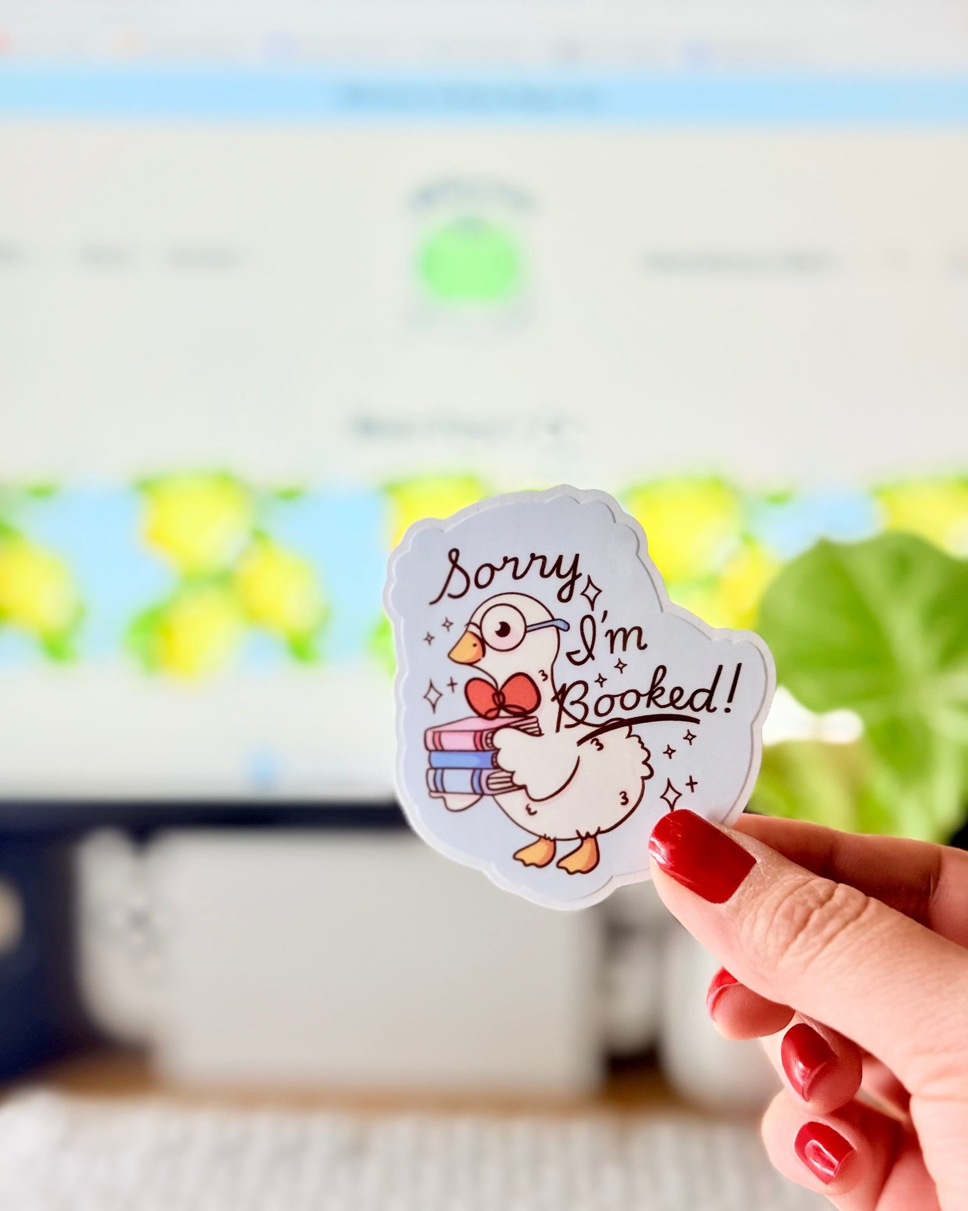 Adorable I'm Booked Duck Sticker. Hand holding a pastel vinyl sticker of a white goose wearing glasses and holding a stack of books with the text “Sorry I’m Booked!” in front of a computer keyboard and plant.