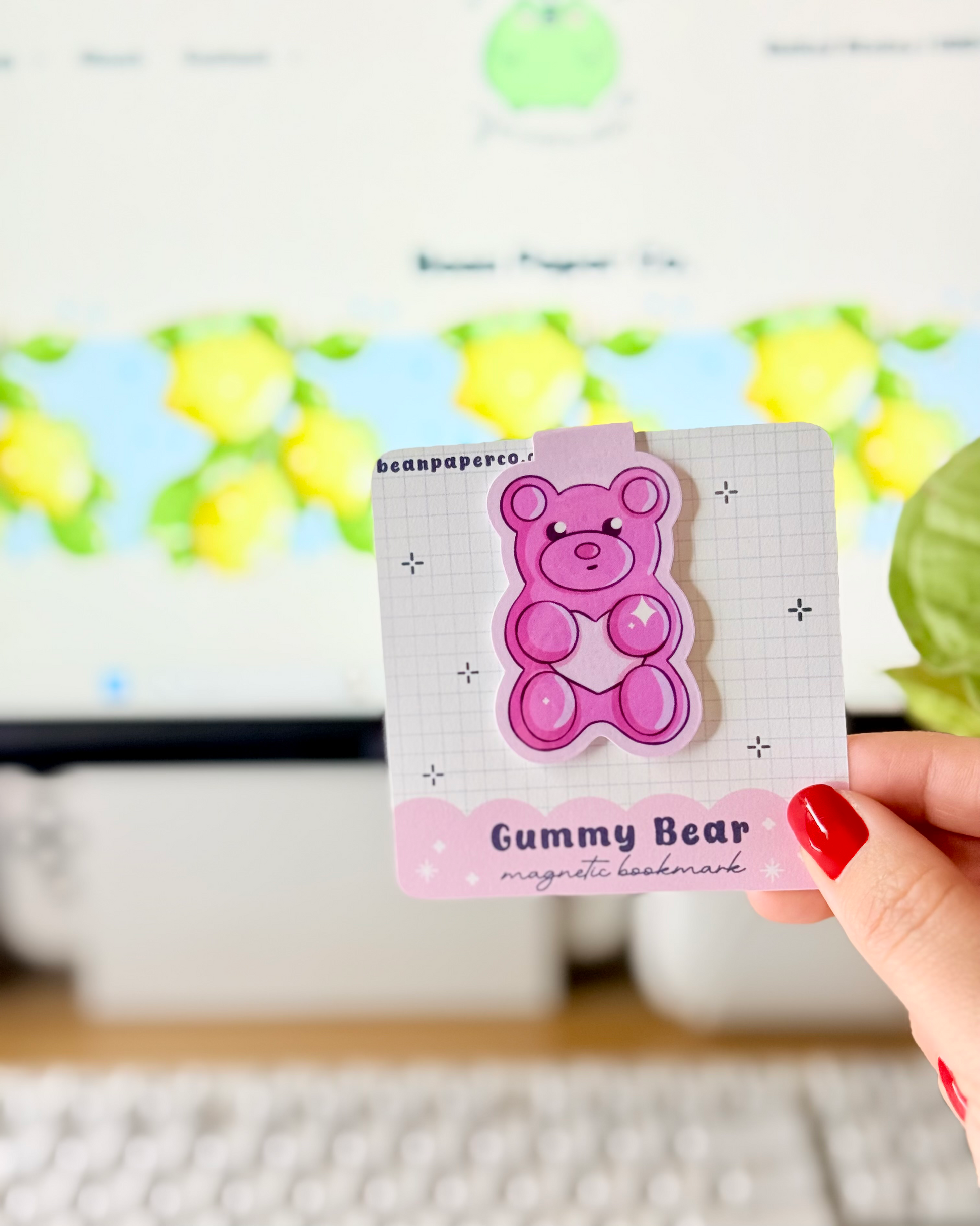 Red & Pink Gummy Bear Magnetic Bookmarks held in hand against blurred Bean Paper Co. website background.
