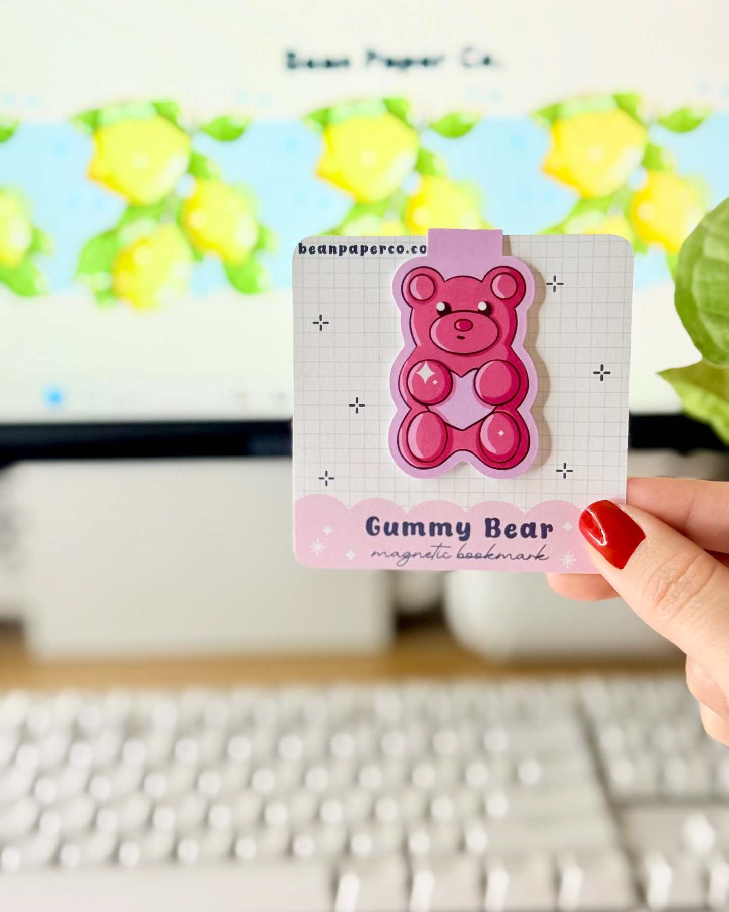 Red & Pink Gummy Bear Magnetic Bookmarks held in hand against blurred Bean Paper Co. website background.