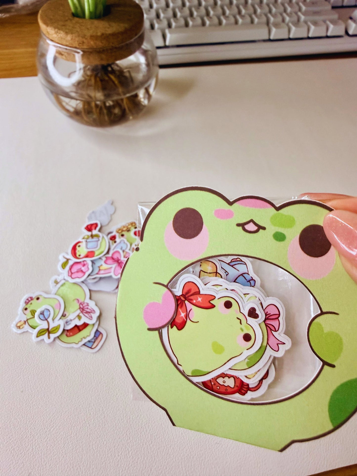 “Frog-shaped packaging holding small die-cut frog sticker flakes on a desk setup.”